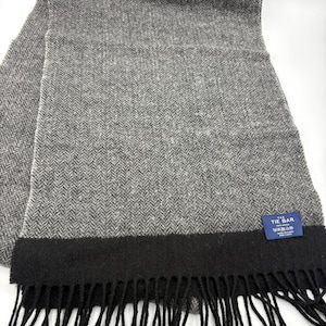 May include: A gray herringbone-patterned scarf with a black border and fringe. The scarf has a small blue tag with white text. The scarf is made of a soft, woven material.