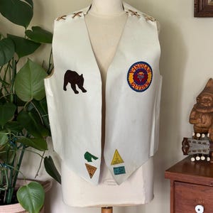 May include: A vintage white vest with a V-neck design, featuring a brown bear patch and several other colorful patches. The vest has decorative stitching and a circular patch that reads "Indian Guides".