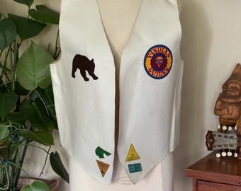 May include: A vintage white vest with a V-neck design, featuring a brown bear patch and several other colorful patches. The vest has decorative stitching and a circular patch that reads "Indian Guides".