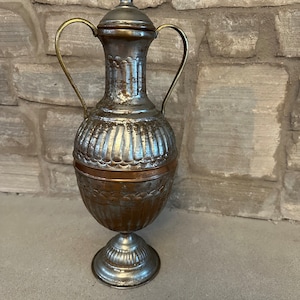 May include: A silver and copper colored metal vase with two handles and a decorative design. The vase has a round body and a pedestal base.