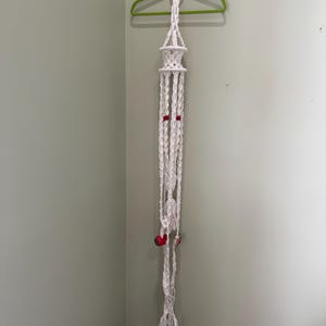 May include: A white macrame plant hanger with red accents and a fluffy tassel at the bottom. The plant holder is suspended from a green hanger with a red ring at the top. The macrame design features intricate knots and a decorative pattern.