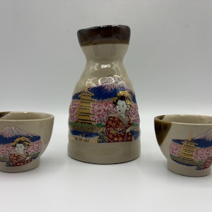 May include: A ceramic sake set consisting of one carafe and two small cups. Each piece features a beige base with a dark brown rim and a colorful graphic depicting a geisha, a pagoda, cherry blossoms, and Mount Fuji. The text MADE IN JAPAN is visible.