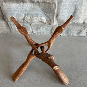 May include: A handcrafted wooden stand with a unique, intertwined branch design. The stand is made of light brown wood and features four upward-pointing branches that meet in the center. The stand is designed to hold a small object.
