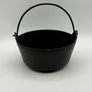 May include: A black cast iron cauldron with a curved handle. The cauldron is round with a slightly flared rim. The handle is made of the same material as the cauldron and is attached to the sides.