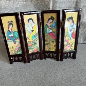 May include: A four-panel folding screen with illustrations of women in traditional Asian attire. The frames are dark brown, and the artwork features a yellow background with floral and plant motifs. Each panel depicts a different woman in a unique pose.