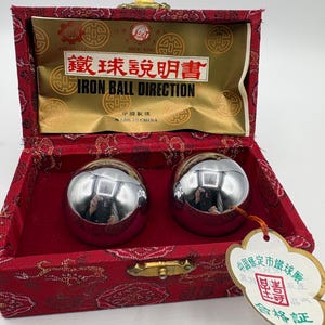 May include: A set of two shiny, chrome-like iron balls inside a red, patterned box. The box has a gold clasp and a gold-colored paper with Chinese text and the words "IRON BALL DIRECTION". A tag with Chinese text hangs from the box.