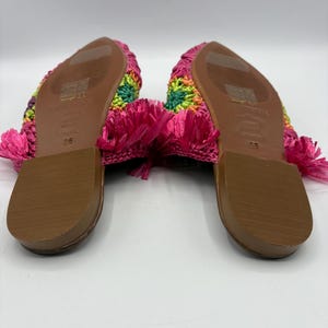 May include: A pair of colorful crochet sandals. The sandals feature a rainbow pattern on the top and a bright pink fringe. The soles are brown. The sandals are size 36.