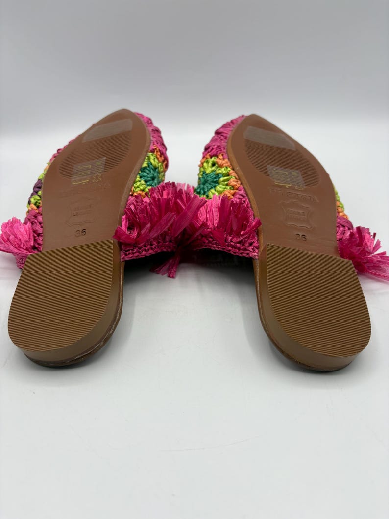 May include: A pair of colorful crochet sandals. The sandals feature a rainbow pattern on the top and a bright pink fringe. The soles are brown. The sandals are size 36.