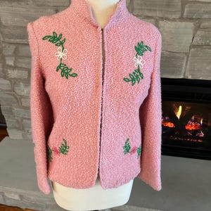 May include: A light pink jacket with a textured weave, featuring embroidered floral designs in white, green, and pink. The jacket has a stand-up collar and a full-length zipper. The jacket is displayed on a mannequin.