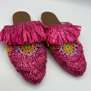 May include: A pair of bright pink raffia slip-on sandals. The sandals feature a woven design with a colorful floral pattern in the center. The top of the sandals have a fringe detail. The soles are brown.