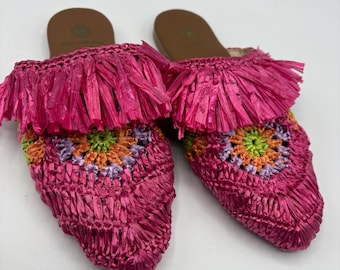May include: A pair of bright pink raffia slip-on sandals. The sandals feature a woven design with a colorful floral pattern in the center. The top of the sandals have a fringe detail. The soles are brown.