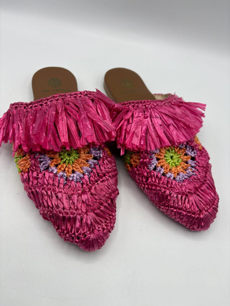 May include: A pair of bright pink raffia slip-on sandals. The sandals feature a woven design with a colorful floral pattern in the center. The top of the sandals have a fringe detail. The soles are brown.