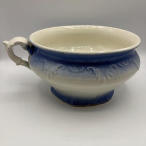 May include: A white and blue ceramic bowl with a handle. The bowl has a wide rim and a footed base. The exterior is decorated with a blue ombre effect and embossed floral designs. The handle is white and ornate.