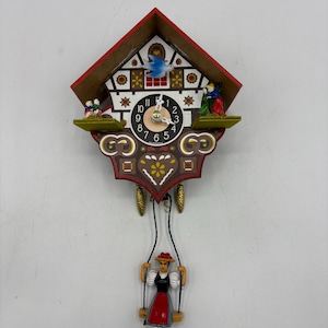 May include: A decorative cuckoo clock with a red roof and a brown wooden body. The clock face is black with white numbers. A small figure of a woman in a red dress is swinging below the clock. Two pinecone weights hang from the bottom.