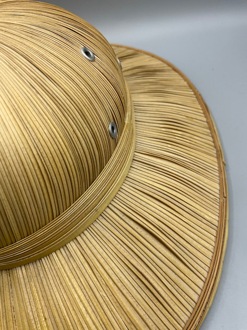 Puede incluir: Un sombrero de paja tejido con un ala ancha y una corona redonda. El sombrero tiene dos ojales de metal en la corona.