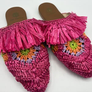 May include: A pair of bright pink raffia slip-on sandals. The sandals feature a fringed upper and a crocheted design with green, orange, and purple accents. The soles are brown with a logo.