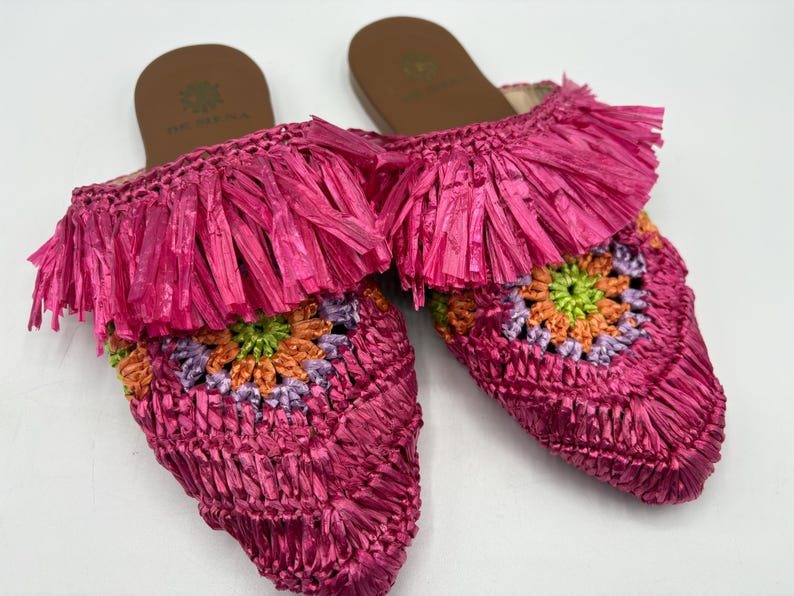 May include: A pair of bright pink raffia slip-on sandals. The sandals feature a fringed upper and a crocheted design with green, orange, and purple accents. The soles are brown with a logo.
