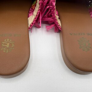 May include: Close-up of two brown leather sandals with the words "DE SIENA" and a floral emblem embossed in gold. The sandals have a woven pink and floral design on the straps, with pink fringe.