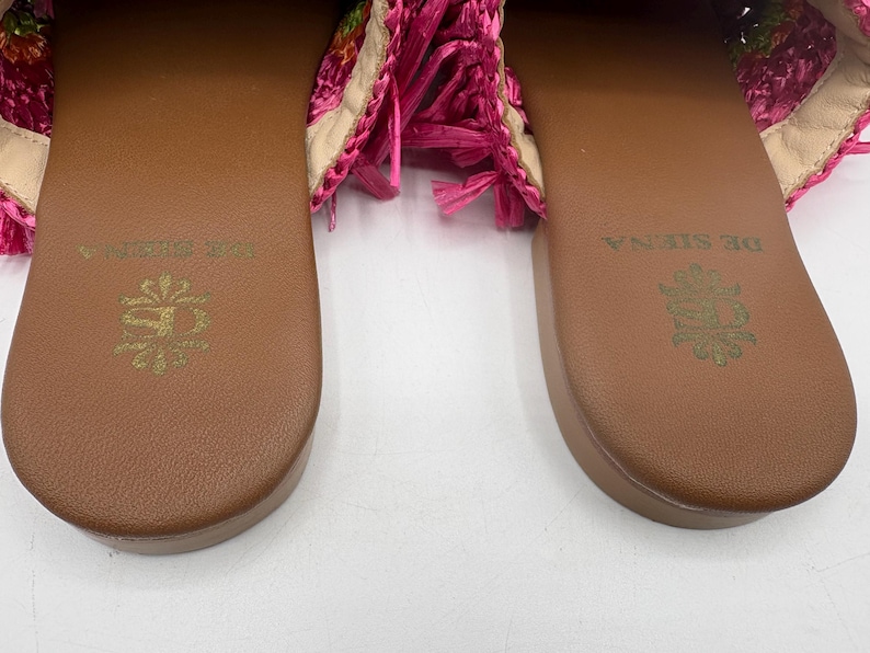 May include: Close-up of two brown leather sandals with the words "DE SIENA" and a floral emblem embossed in gold. The sandals have a woven pink and floral design on the straps, with pink fringe.