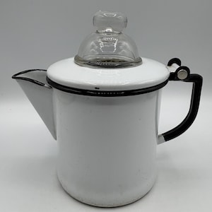 May include: White enamel coffee pot with a black handle and trim. The pot has a glass lid and a spout for pouring. A classic kitchenware item.