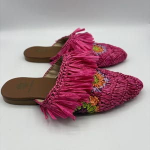 May include: A pair of pink woven raffia slides with fringe detailing. The slides feature a brown sole and a floral design in shades of orange, green, and purple on the top. The shoes are designed for casual wear.