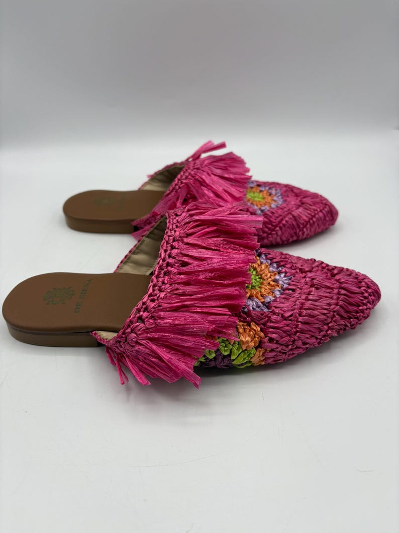 May include: A pair of pink woven raffia slides with fringe detailing. The slides feature a brown sole and a floral design in shades of orange, green, and purple on the top. The shoes are designed for casual wear.