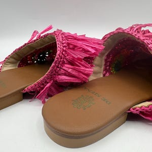May include: A pair of bright pink raffia slides. The slides feature a woven design with fringe detailing and a brown leather sole. The brand name "DE SIENA" is embossed on the footbed. The shoes are designed for casual wear.