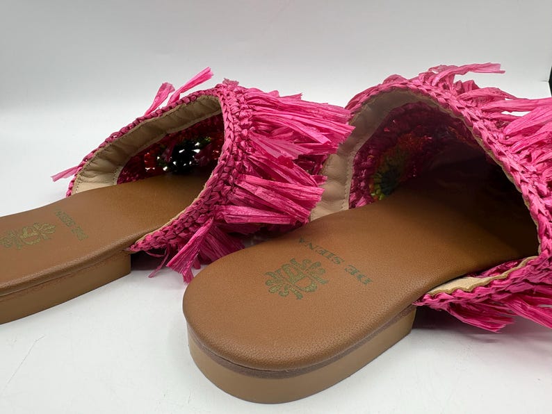May include: A pair of bright pink raffia slides. The slides feature a woven design with fringe detailing and a brown leather sole. The brand name "DE SIENA" is embossed on the footbed. The shoes are designed for casual wear.