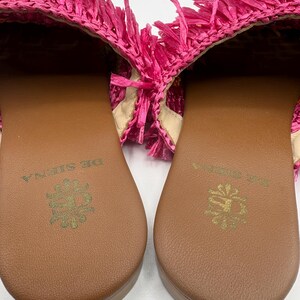 May include: A pair of bright pink raffia sandals. The sandals have a brown sole with a gold emblem and the words "DE SIENA" printed on the sole. The upper part of the sandal is made of pink raffia with fringe.