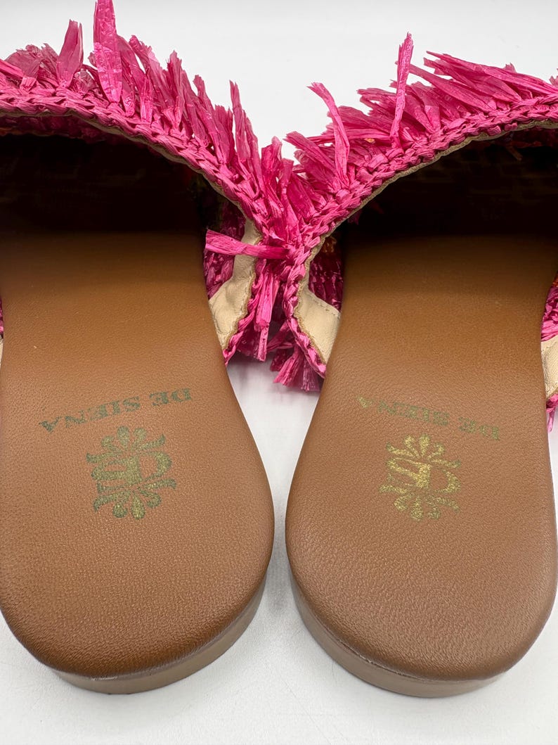 May include: A pair of bright pink raffia sandals. The sandals have a brown sole with a gold emblem and the words "DE SIENA" printed on the sole. The upper part of the sandal is made of pink raffia with fringe.