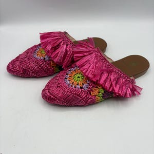 May include: A pair of pink raffia slip-on sandals. The sandals feature a woven design with a floral pattern in shades of orange, green, and purple. Each sandal has a pink fringe detail at the top.