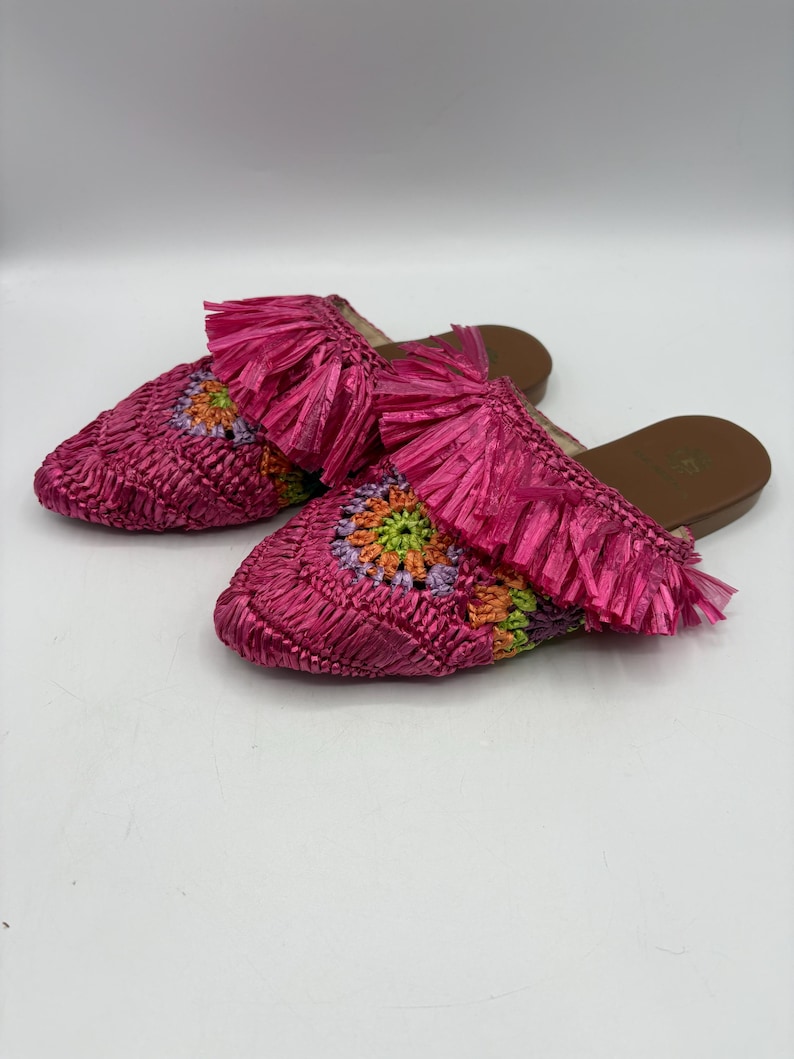 May include: A pair of pink raffia slip-on sandals. The sandals feature a woven design with a floral pattern in shades of orange, green, and purple. Each sandal has a pink fringe detail at the top.