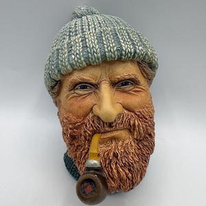 May include: A detailed sculpture of a man's face, wearing a blue and gray knit cap and a full, reddish-brown beard. The man is smoking a pipe with a yellow stem. The sculpture has a weathered appearance, with visible wrinkles and blue eyes.