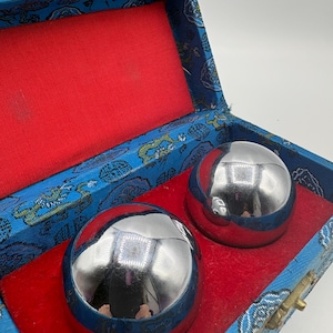 May include: Two polished silver metal balls rest inside a blue and red decorative box. The box has a blue fabric exterior with a floral pattern and a red velvet interior. The box is closed with a gold-colored clasp.