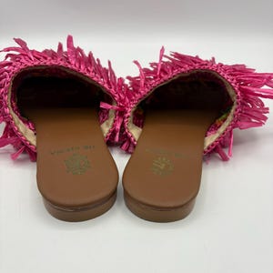 May include: A pair of open-toe sandals with a brown sole and a bright pink fringe detail. The sandals have a woven texture and a floral pattern on the inner sole. The brand name "DE SIENA" is printed on the sole.