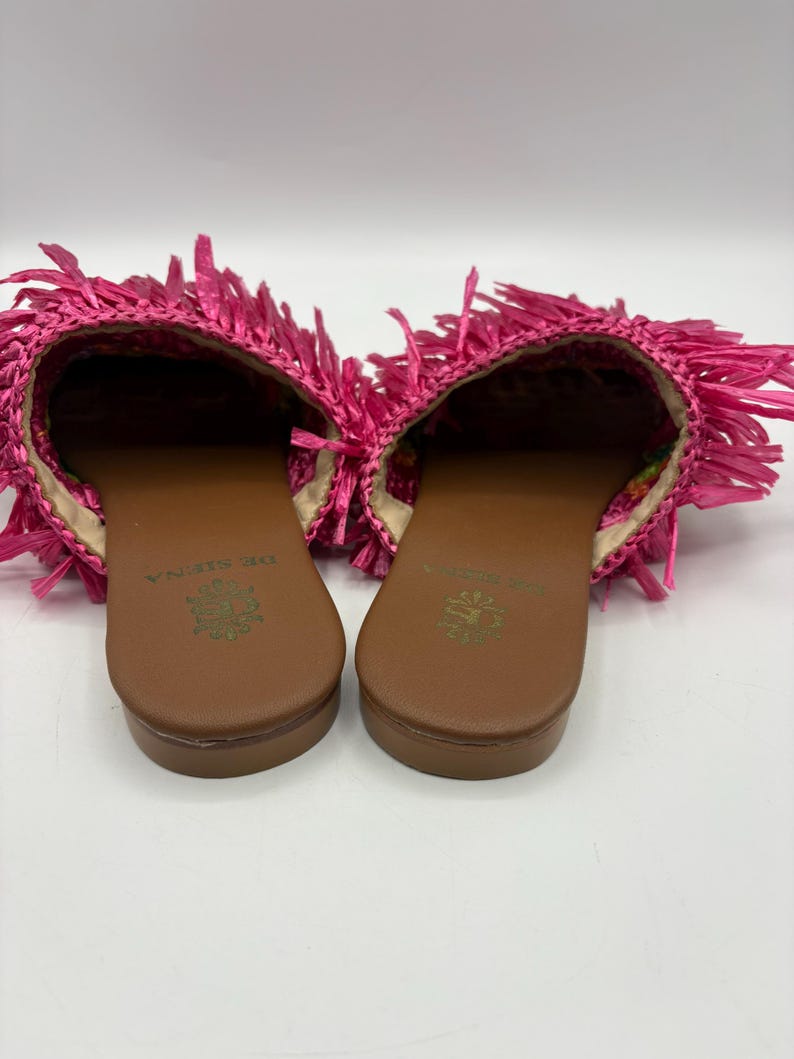 May include: A pair of open-toe sandals with a brown sole and a bright pink fringe detail. The sandals have a woven texture and a floral pattern on the inner sole. The brand name "DE SIENA" is printed on the sole.