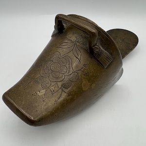 May include: A brass shoe shaped container with a floral design and a handle. The container is made of a dark brown metal and has a worn finish.