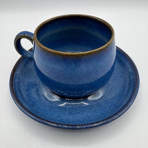May include: A blue ceramic teacup and saucer set. The cup has a rounded shape with a handle and sits on a matching saucer. The set has a glossy finish with a darker brown rim.