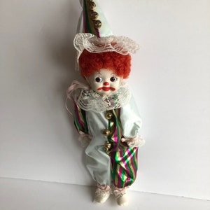 May include: A porcelain clown doll with a red curly wig, a white ruffled collar, and a matching hat. The doll's outfit is a light blue satin with green and pink striped accents and gold bells.