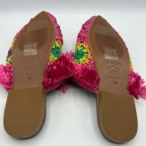 May include: A pair of colorful, woven mules. The soles are brown with the size 36 printed on them. The uppers are a mix of bright colors, including pink, green, orange, and purple. The heels have a pink fringe.