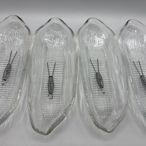 May include: Four clear glass corn-shaped dishes with textured exteriors. Each dish contains a silver-colored corn cob holder. The dishes are arranged side by side, showcasing their design and intended use for serving corn on the cob.