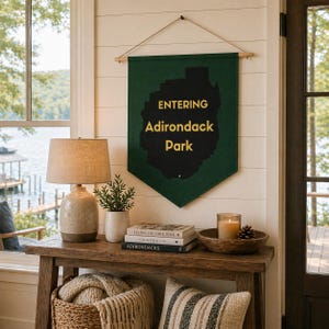 May include: A green banner with the text "ENTERING Adirondack Park" hangs on a white wall. A wooden table displays a lamp, plant, books, candle, and a basket with a blanket. A window overlooks a lake.