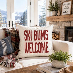 Può includere: Un cuscino quadrato color crema con le parole "SKI BUMS WELCOME" in lettere rosse. Il cuscino è su un divano bianco con una coperta a quadri e un cuscino a righe blu. Una piccola pianta in vaso è su un tavolino di legno.