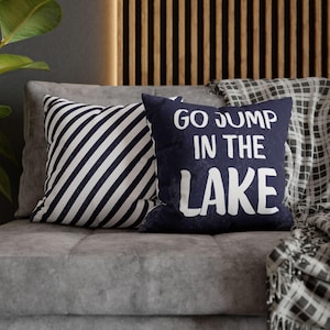 May include: Two throw pillows on a gray couch. One pillow is navy blue with white text that reads "Go Jump in the Lake". The other pillow is black and white striped.