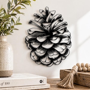 May include: Black and white metal pine cone wall art. The decorative piece is mounted on a white wall. The pine cone is a detailed, stylized illustration. The image also shows a vase with greenery, books, and wooden beads.