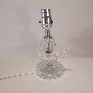May include: A clear glass lamp base featuring a textured, faceted design with a round, scalloped bottom and a small handle on the side. The lamp includes a silver-toned metal socket at the top and a white electrical cord extending from the base.