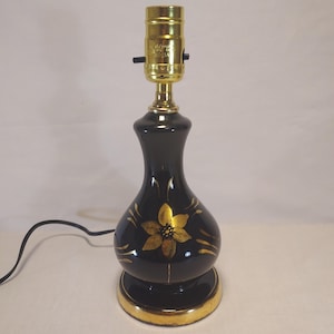May include: A black ceramic table lamp base with a gold floral design and a gold-colored metal socket. The lamp features a rounded, bulbous body, a circular base with a gold rim, and a black electrical cord extending from the side of the base.
