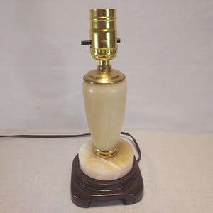 May include: A table lamp featuring a smooth, cream-colored stone body with subtle veining, set upon a circular stone base and a dark brown square wooden stand. The lamp includes a polished gold-tone metal socket assembly with a black switch.