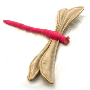 Puede incluir: Un juguete de libélula de tela rosa y beige con detalles dorados. La libélula tiene un cuerpo largo y delgado y dos pares de alas.