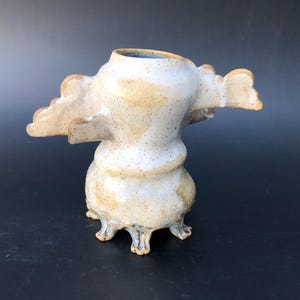 May include: A ceramic vessel with a speckled off-white and tan glaze, featuring a wide, rounded base supported by small, claw-like feet. The body narrows toward the center and flares out at the top with two symmetrical, wing-like protrusions.
